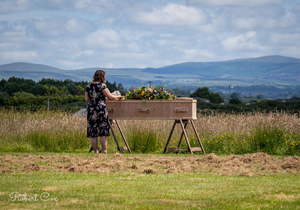 Pembrokeshire-Heart of Pembrokeshire-Ecological Coffins-Pembs Petals-11 Celebrant by a wooden coffin at Pembrokeshire Natural Burial Ground